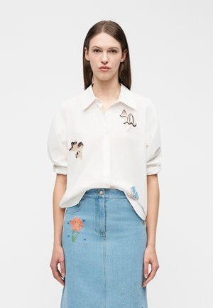 Kvinde iført en hvid skjorte med svampebroderi og en blå jeansnederdel med blomsterbroderi, stående mod en ensfarvet baggrund.