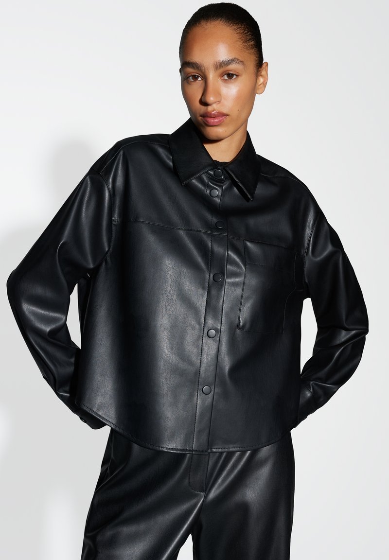 Camicia in finta pelle nera con colletto, bottoni a scatto e taschino sul petto. Presenta una texture liscia e un design leggermente corto.