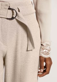 Beige hoch taillierte Hose mit einem Stoffgürtel, aus strukturiertem Material. Aufwendige Spitzenverzierung am Ärmel eines hellen Oberteils.