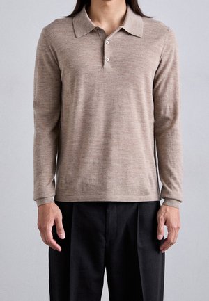 Personne portant un polo à manches longues beige avec deux boutons et un pantalon noir, debout devant un fond clair uni.