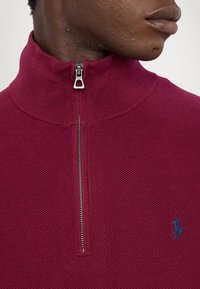 Gros plan sur une personne portant un pull zippé texturé couleur bordeaux avec un col relevé et un logo joueur de polo bleu marine sur la poitrine.