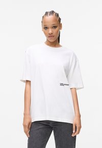 Hvid bomulds T-shirt med korte ærmer og rund hals, med et sort logo på venstre side. Sammen med mørkegrå denimjeans.