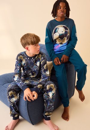 Zwei barfüßige Kinder in passenden Pyjamas mit Fußballmotiv, eines sitzt auf einem blauen Kissen, das andere auf dessen Armlehne.