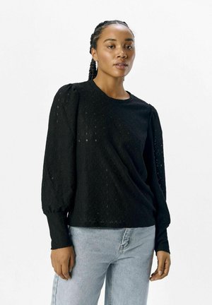 Pull noir texturé avec de longues manches bouffantes, encolure ronde et motif subtil en losanges. Associé à un jean en denim bleu clair.