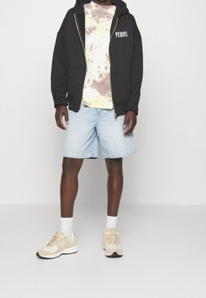 Person wearing black zip-up hoodie with "PEQUS" text, tie-dye t-shirt, light blue denim shorts, white socks, and beige sneakers.