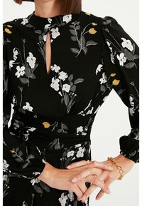 Zwart jurkje met bloemenprint in wit, grijs en geel. Sleutelgat halslijn, pofmouwen en een getailleerd detail. Gouden armband.