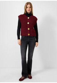 Bordo adīts veste ar teksturētu rakstu, ar četriem zeltainiem pogām un divām priekšējām kabatām, vilkta virs melna garām piedurknēm tee un džinsiem.