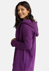 Długi purpurowy hoodie wykonany z miękkiego materiału, z zamkiem błyskawicznym z przodu, bocznymi kieszeniami i dużym kapturem. Design podkreśla komfort i przytulność.