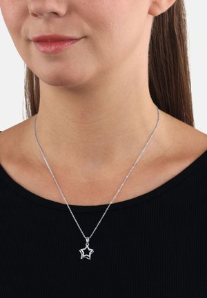 Collana con pendente a forma di stella in argento, con una catena delicata, caratterizzata da un design a intaglio e una texture sottile, indossata su una maglia nera.