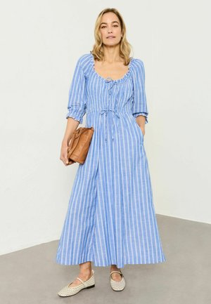 Frau trägt ein blau-weiß gestreiftes Midikleid mit 3/4-Ärmeln, hält eine braune Handtasche und trägt bequeme beige geflochtene flache Schuhe.