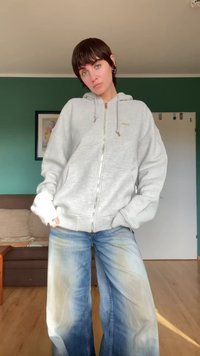 Gray zip-up hoodie with a drawstring hood, paired with light blue wide-leg jeans featuring faded sections. Wood floor and green wall backdrop.