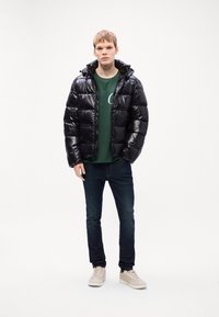 Νεαρός άνδρας που φοράει μια μαύρη φούτερ ζακέτα, πράσινη μπλούζα, σκούρο τζιν και ανοιχτό μπεζ αθλητικά παπούτσια, που στέκεται μπροστά σε απλό λευκό φόντο.