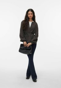 Teksturowany ciemny kardigan z paskiem w talii, zestawiony z białym topem i dzwonowatymi ciemnymi jeansami. Modelka trzyma czarną luźną torebkę.