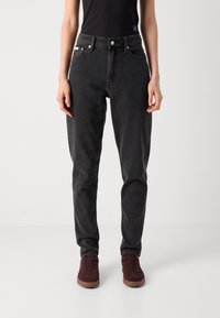 Asmuo, vilkintis aukštai argančiais juodais džinsais ir bordo spalvos sportbačiais, stovi prieš paprastą šviesiai pilką foną.