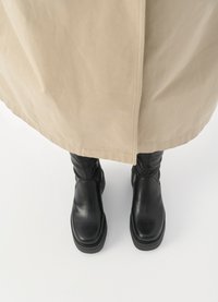 Botas de cuero negras hasta el tobillo con un acabado liso, que presentan una suela gruesa y un diseño elegante, combinadas con una prenda exterior beige de largo hasta la rodilla.