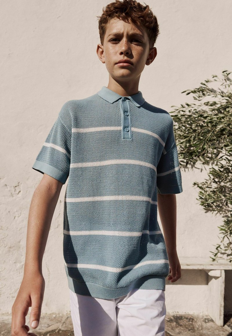 Niño con cabello rizado que lleva una camisa polo de punto a rayas azul y blanca y pantalones blancos, de pie al aire libre junto a una pared blanca y una planta.