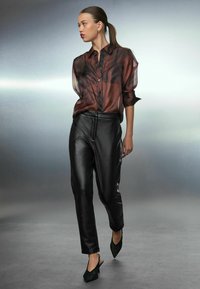 Sheer brown and black patterned blouse, black faux leather pants, and green suede heeled mules worn against a softly lit background.