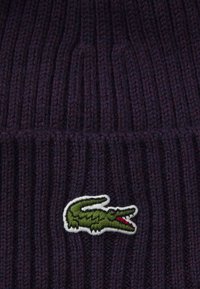 Bonnet en tricot de couleur violet foncé avec un motif côtelé texturé. Il présente un logo de crocodile brodé en vert et blanc à l'avant.