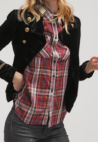 Giacca di velluto nero con bottoni dorati, sovrapposta a una camicia a quadri nei toni rosso, blu e bianco, con colletto classico e chiusura a bottoni.