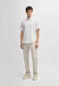 Camicia bianca a maniche corte con bottoni, dalla vestibilità rilassata, abbinata a pantaloni beige chiaro e sneakers color crema. Design minimalista.