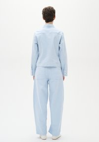 Gai zils džinsas žakets un platā kāju bikses, abas ar gludu tekstūru, pogu akcentiem un aizmugurējām kabatām, skatītās no aizmugures.