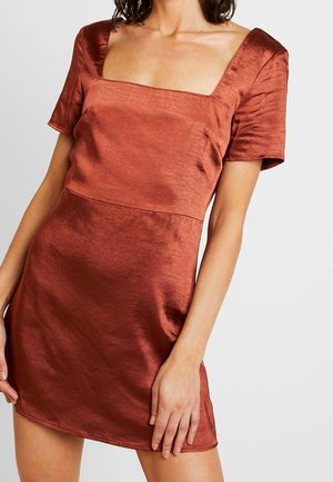 Femme portant une mini-robe en satin couleur rouille à manches courtes avec un col carré, debout devant un fond uni.
