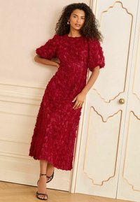 Red dress covered in fabric rose appliqués, A-line silhouette, short puff sleeves, knee-length, paired with strappy black sandals.
