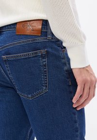 Granatowe dżinsy z pięcioma kieszeniami, z brązową skórzaną naszywką z logo Calvin Klein na pasku i kontrastowym szyciem.