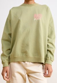 Personne portant un sweat-shirt vert clair avec le texte rose "Always Salty Roxy" sur la poitrine et un pantalon de couleur crème sur un fond blanc.