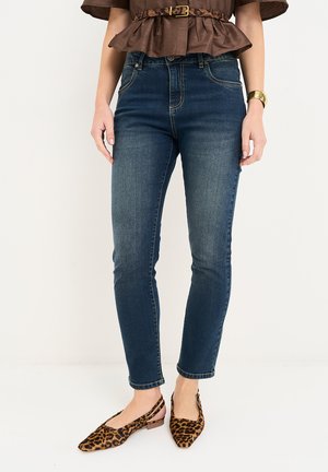 Kvinde iført mørkeblå skinny jeans, brun kortærmet bluse med leopardprint bælte, leopardprint spidse flade sko og guldt armbånd.