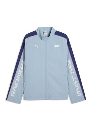 Lyseblå Puma jakke med mørkeblå ærmer, pink piping, "RACING" på venstre ærme, "EST. 1950" på højre ærme og F1-logo på brystet.