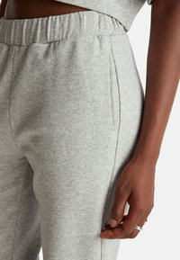 Šedé tepláky z měkké látky s elastickým pasem, vybavené bočními kapsami a jemnou texturou. Detail pasu a ruky.