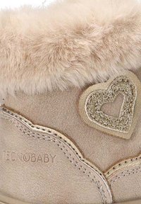 Calzado beige con ribete de piel sintética, con un adorno en forma de corazón y costuras decorativas. Textura suave y forma redondeada.