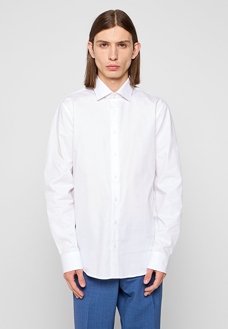 Mann mit glattem braunem Haar, der ein weißes Langarmhemd und blaue Hosen trägt, steht vor einem einfarbigen hellen Hintergrund.