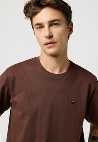 Ruskea puuvillapaita, jossa on pyöreä kaula-aukko ja lyhyet hihat; sisältää pienen mustan logopatchin vasemmassa rinnassa. Tasainen pinta.