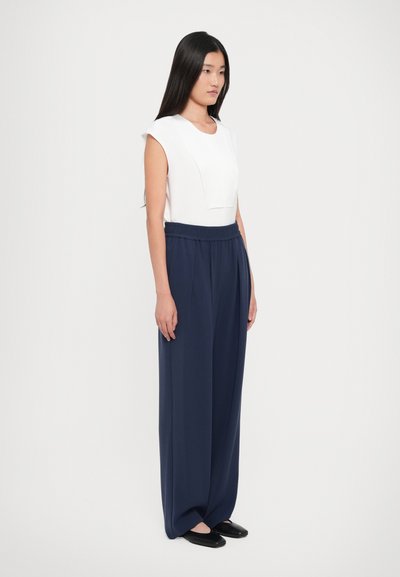 Asian woman standing, wearing sleeveless white top, navy wide-leg trousers, and black flat shoes, against a plain light background.