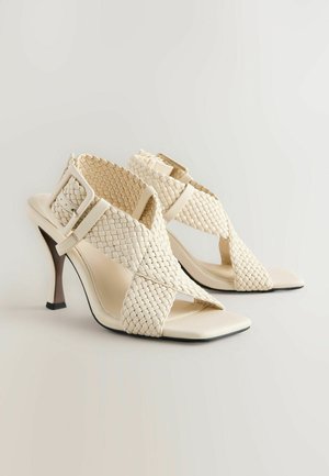 Beige högklackade sandaler med vävd överdel, öppen tådesign, korsade remmar och en slank stilettklack. Har en spänne som accent.