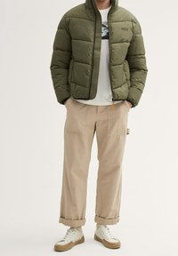 Olivengrøn puffjakke med høj krave, lynlås foran og quiltet design, kombineret med beige bukser med vide ben og hvide sneakers.