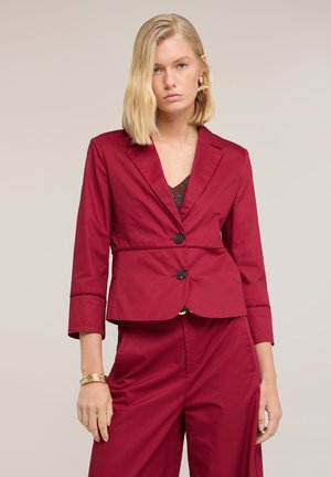 Femme blonde portant un blazer rouge ajusté avec deux boutons et un pantalon taille haute assorti, debout devant un fond uni.