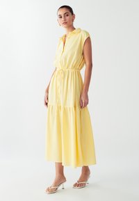 Geltona, be rankovių suknelė su apykakle, sagčių uždarymu ir raišteliu liemeniui. Suknelė turi daugiapakopį sijoną ir lengvą audinį.