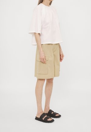 Person iført en løs hvid kortærmet top, beige knælange cargoshorts og sorte sandaler med åben tå, stående mod en ensfarvet baggrund.