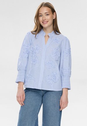Jonge vrouw die een blauw-wit gestreept blouse draagt met bloemrijke borduursels en ballonmouwen, gecombineerd met een blauwe jeans, licht glimlachend.