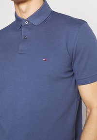 Man som bär en blå kortärmad polo med två knappar och en liten röd, vit och blå rektangulär logotyp på vänster bröst.