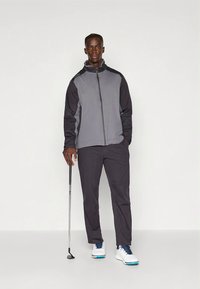 Homme se tenant contre un fond blanc, portant une veste grise et noire, un pantalon noir, des chaussures de golf blanches, tenant un club de golf dans la main droite.