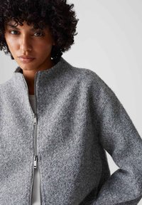 Giacca grigia con zip realizzata in tessuto testurizzato, caratterizzata da un colletto alto e dettagli in metallo argentato, con una vestibilità ampia e maniche lunghe.