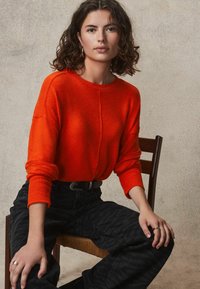 Oranger Wollpullover mit gerippten Bündchen und Rundhalsausschnitt, kombiniert mit schwarzen strukturierten Jeans. Modell sitzt auf einem Holstuhl vor einem neutralen Hintergrund.