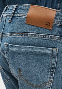 Κοντινό πλάνο μπλε τζιν με ορατή πίσω τσέπη και ραμμένο καφέ δερμάτινο λογότυπο στη ζώνη μέσης.