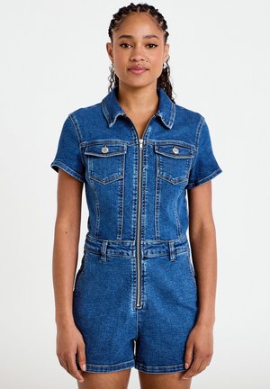 Kvinde med fletninger iført kortærmet blå denimkortdragt med frontlynlås, brystlommer og bæltestropper, stående mod ensfarvet baggrund.
