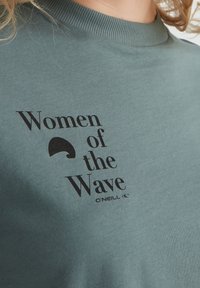 Merenvihreä puuvillainen t-paita, jossa on pyöreä pääntie ja mustalla painettu teksti "Women of the Wave" sekä aaltoaiheinen grafiikkadetaili.