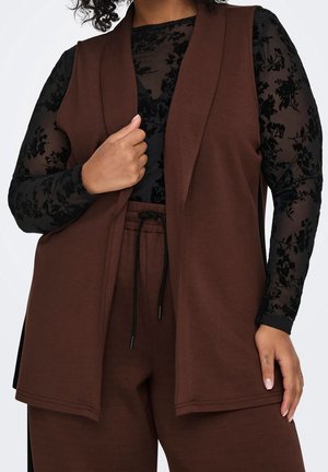 Kvinde iført brun ærmeløs vest og bukser med sort transparent blomstret langærmet top, hånd holder på vestens revers.